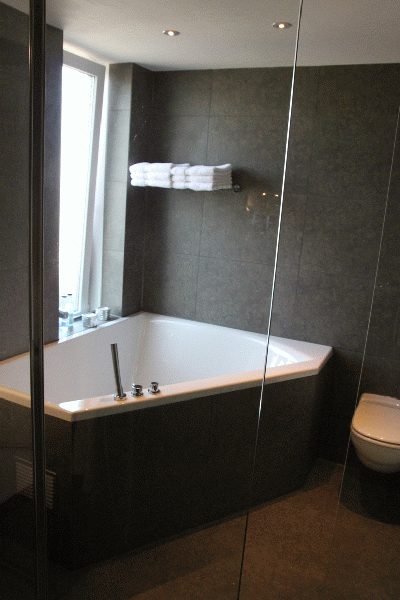 Gerenoveerde badkamer met antracietgrijze wandtegels, hoekbad en glazen douchewand.